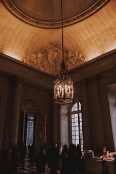 An ornate interior space with a grand chandelier hanging from a detailed, decorative ceiling. The room showcases intricate moldings and classical architecture, with tall windows allowing natural light to filter through. A group of people is gathered around, possibly attending an event or exploring the location.