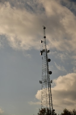 A tall communication tower stands against a sky partially filled with soft, scattered clouds. The tower is equipped with various antennas and dishes, silhouetted against the lighter background of the sky.
