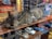 A tabby cat is lying on a wooden shelf inside a store, surrounded by various objects. Next to the cat, there are miniature model cars in different colors including white, blue, and red. A framed picture or sign with the word 'Wasser' in gold letters is visible behind the cat. Reflections of the street are seen on the glass, indicating this is a window display.