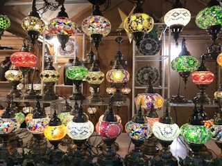 A collection of ornate hanging lamps is displayed, featuring colorful stained glass designs and intricate metalwork. The lamps exhibit various vibrant patterns, including mosaics and geometric shapes, creating a rich visual tapestry.