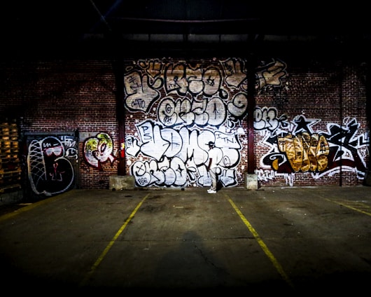A brick wall covered with various graffiti and street art designs, showcasing different styles and colors. In front of the wall, a paved area with faded yellow parking or boundary lines is visible. The scene is dimly lit, suggesting an urban environment or warehouse setting.