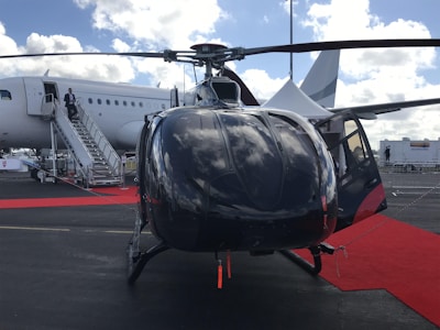 A helicopter is positioned in the foreground on a tarmac with a red carpet leading up to it. In the background, a large airplane is visible with someone descending its stairs. The sky is partly cloudy with light reflecting off the surfaces of the helicopter.