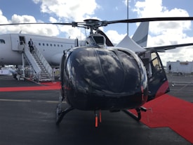 A helicopter is positioned in the foreground on a tarmac with a red carpet leading up to it. In the background, a large airplane is visible with someone descending its stairs. The sky is partly cloudy with light reflecting off the surfaces of the helicopter.