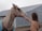 A light-colored horse wearing a blue blanket is being gently touched by a person with long hair wearing a brown sweater. The interaction takes place near a wooden fence, with a large barn or shed in the background.