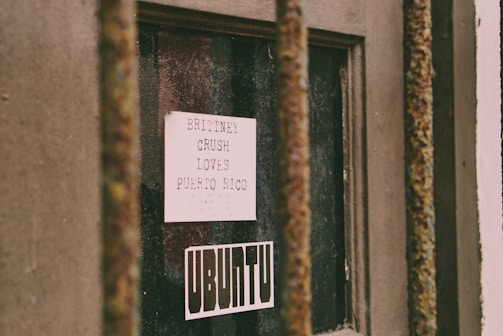 A close-up view of a weathered window protected by rusted metal bars. Inside the window is a white piece of paper with typed text that reads 'BRITTNEY CRUSH LOVES PUERTO RICO.' Below the paper is a sticker with the word 'UBUNTU' in bold typography. The scene suggests a blend of personal expression and urban decay.