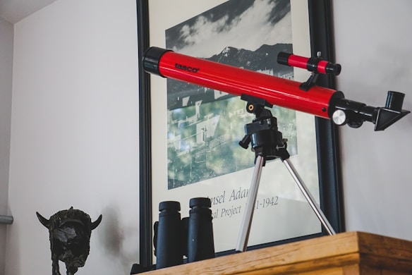 A red telescope mounted on a tripod is placed on a wooden shelf. Below the shelf are binoculars. Behind the telescope, there is a framed black-and-white photograph with text 'Ansel Adams' and '1941-1942' visible. To the left, a dark sculpture resembling an animal head is positioned under the shelf.