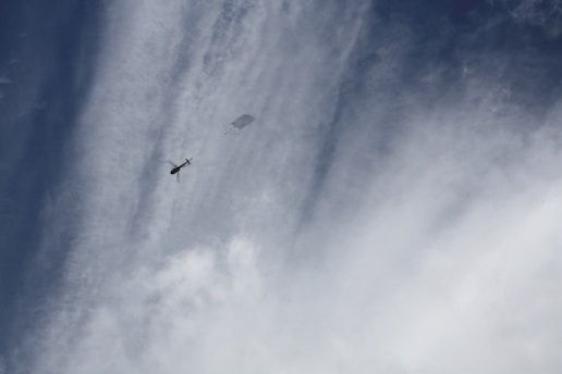 A helicopter is flying high in the sky, towing a rectangular object. The sky is filled with scattered clouds and streaks of white, providing a dynamic and slightly dramatic background.