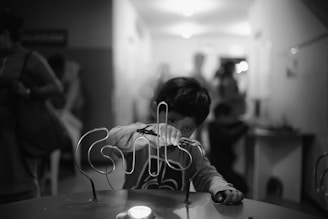 A child focuses intently on maneuvering a metal wire loop skill game. The background is softly blurred with other people in the distance.
