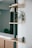 A minimalist interior space with wooden shelves mounted on a wall. A small green plant with long leaves is placed on one shelf, and a small decorative figurine sits on another. The background is softened, emphasizing the clean lines and natural textures of the furniture.