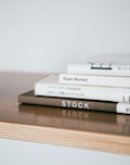 A stack of neatly arranged hardcover books sits on a light wooden surface. The books have minimalist designs with some text visible on the spines, including titles like 'Super Normal' and 'STOCK.' The overall composition is simple and aesthetically pleasing.
