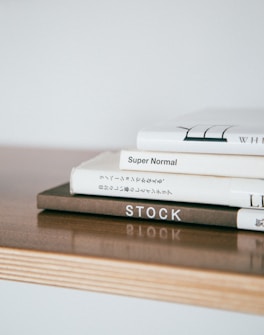A stack of neatly arranged hardcover books sits on a light wooden surface. The books have minimalist designs with some text visible on the spines, including titles like 'Super Normal' and 'STOCK.' The overall composition is simple and aesthetically pleasing.