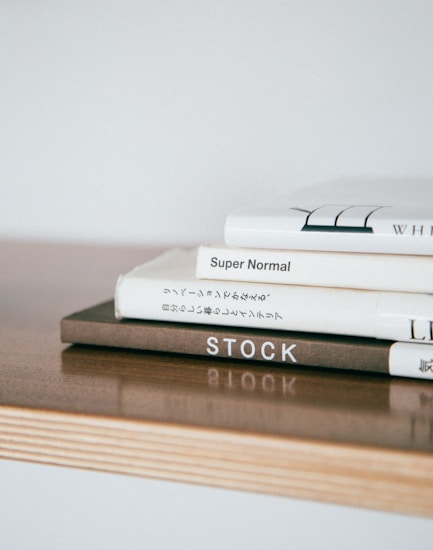A stack of neatly arranged hardcover books sits on a light wooden surface. The books have minimalist designs with some text visible on the spines, including titles like 'Super Normal' and 'STOCK.' The overall composition is simple and aesthetically pleasing.