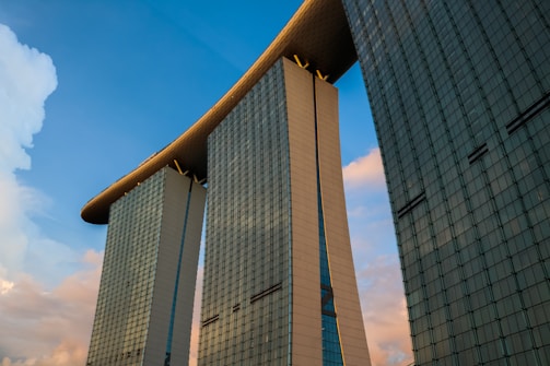 Three towering skyscrapers connected at the top by a unique and massive horizontal structure against a backdrop of blue sky with some clouds. The buildings have a modern architectural design with glass facades reflecting the light.