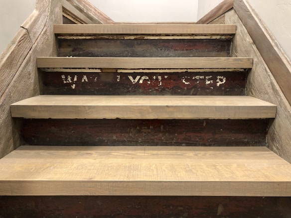 A set of worn, wooden stairs with a rustic appearance. The dark brown steps have visible grain patterns, and on the riser of one step, partially visible, faded white text reads 'WATCH YOUR STEP'. The stairs are bordered by rough-hewn wooden rails, contributing to an aged, industrial vibe.
