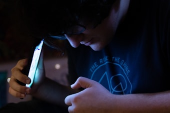 A person is intently looking at a smartphone held close to their face, casting a soft glow on their features. The background is dimly lit, emphasizing the contrast between the darkness and the bright screen.
