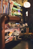 A bartender is behind the counter, making a drink with various ingredients and tools laid out in front of them. There are several bottles on the shelves in the background, and the scene is warmly lit with a cozy ambiance.