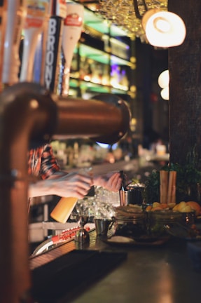 A bartender is behind the counter, making a drink with various ingredients and tools laid out in front of them. There are several bottles on the shelves in the background, and the scene is warmly lit with a cozy ambiance.