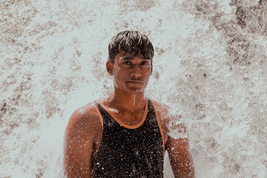 A person is standing in front of a forceful stream of water, likely from a waterfall. The individual is wearing a black tank top and appears to be slightly wet from the splashing water. The background is filled with the white, foamy water, giving a sense of movement and energy.