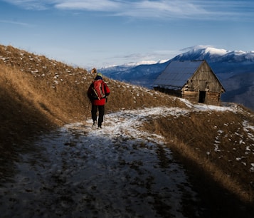 A person dressed in winter clothing, carrying a backpack and walking stick, is hiking along a snowy path on a hillside. The sun casts long shadows, and in the distance, there is a wooden cabin with a slanted roof. Snow-capped mountains are visible under a clear blue sky.