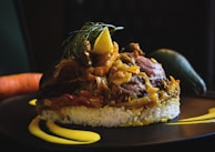 A finely plated dish with a base of white rice topped with a savory mixture of meat and vegetables, garnished with a lemon slice and fresh herbs. The plate includes artistic streaks of yellow sauce, and whole vegetables like a carrot and an avocado rest in the background.