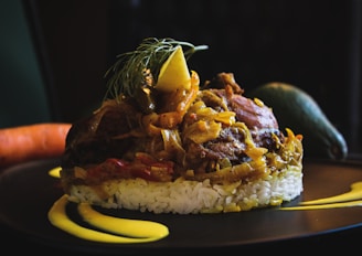A finely plated dish with a base of white rice topped with a savory mixture of meat and vegetables, garnished with a lemon slice and fresh herbs. The plate includes artistic streaks of yellow sauce, and whole vegetables like a carrot and an avocado rest in the background.
