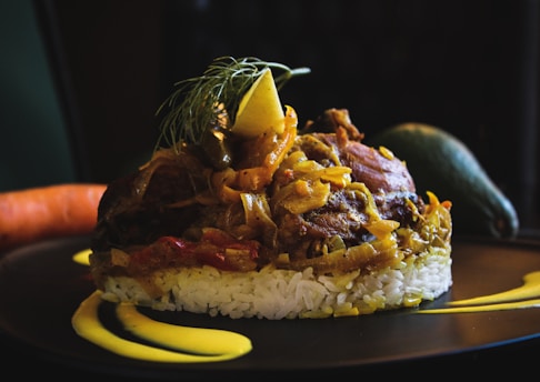 A finely plated dish with a base of white rice topped with a savory mixture of meat and vegetables, garnished with a lemon slice and fresh herbs. The plate includes artistic streaks of yellow sauce, and whole vegetables like a carrot and an avocado rest in the background.