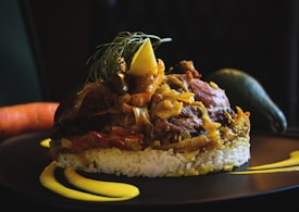 A finely plated dish with a base of white rice topped with a savory mixture of meat and vegetables, garnished with a lemon slice and fresh herbs. The plate includes artistic streaks of yellow sauce, and whole vegetables like a carrot and an avocado rest in the background.
