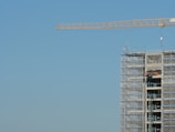 A construction site features a tall building in the process of being built, surrounded by scaffolding. A large crane extends across the clear blue sky, indicating ongoing construction work.
