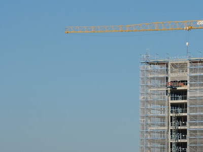 A construction site features a tall building in the process of being built, surrounded by scaffolding. A large crane extends across the clear blue sky, indicating ongoing construction work.