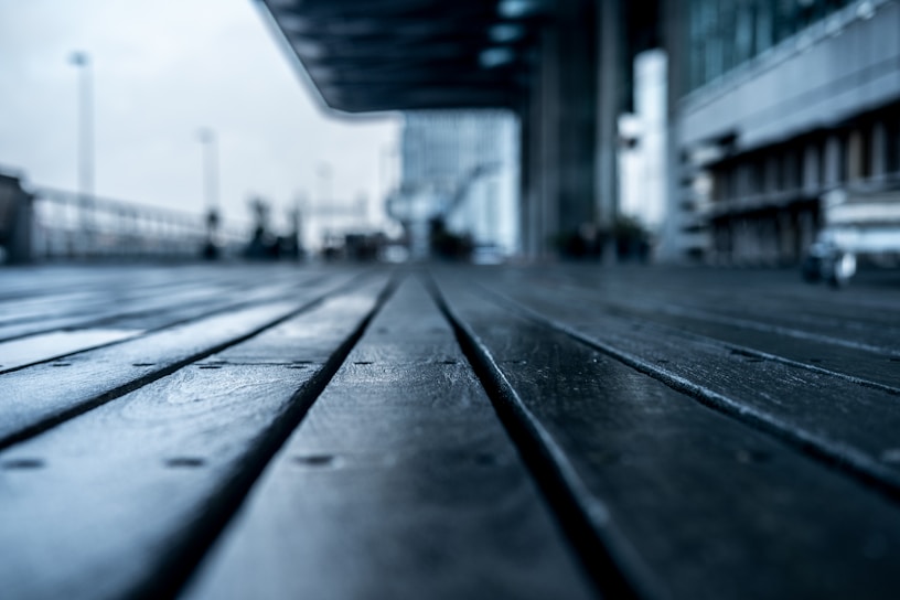 A wooden deck extends into the distance, with a low angle perspective that emphasizes the texture of the wooden planks. In the background, an out-of-focus urban environment features modern buildings and structures. The image is slightly desaturated, with a cool color tone.