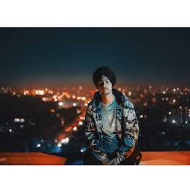 A person wearing a turban and a camouflage jacket is sitting on a ledge, with a cityscape in the background. The city lights create a bokeh effect, giving the scene a vibrant and energetic feel.