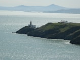 A picturesque coastal scene features a lighthouse on a rocky cliff overlooking a vast body of shimmering water. Nearby, a white building rests on the grassy cliff. In the background, distant hills form a serene silhouette against the horizon.