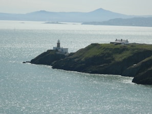 A picturesque coastal scene features a lighthouse on a rocky cliff overlooking a vast body of shimmering water. Nearby, a white building rests on the grassy cliff. In the background, distant hills form a serene silhouette against the horizon.