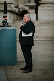 A man in a dark suit and tie, with white hair and glasses, stands on an urban street. He is holding a newspaper under his arm and smoking a pipe. The background features stone buildings and steps.