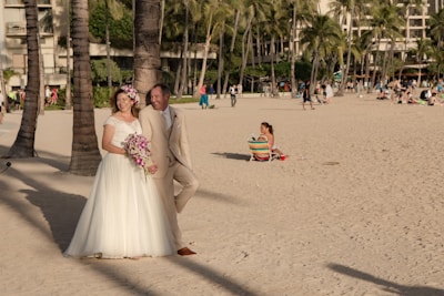 A couple is dressed in wedding attire on a sandy beach with palm trees and buildings in the background. The bride is wearing a white gown and floral crown, holding a bouquet of pink and purple flowers. The groom is in a beige suit. Several people are enjoying the beach in the background.