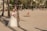 A couple is dressed in wedding attire on a sandy beach with palm trees and buildings in the background. The bride is wearing a white gown and floral crown, holding a bouquet of pink and purple flowers. The groom is in a beige suit. Several people are enjoying the beach in the background.