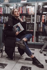 A person with light hair and a contemplative expression sits on a chair in a bookstore, holding a book titled 'Bosch'. The individual is dressed in a black coat, jeans, and dark boots, and appears relaxed. Behind them, a bookshelf is filled with a variety of books, and a shopping cart is partially visible to the right.