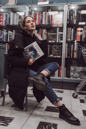 A person with light hair and a contemplative expression sits on a chair in a bookstore, holding a book titled 'Bosch'. The individual is dressed in a black coat, jeans, and dark boots, and appears relaxed. Behind them, a bookshelf is filled with a variety of books, and a shopping cart is partially visible to the right.