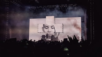 A concert stage is lit up with an impressive visual display featuring a large screen showing a black and white portrait of a person's face. Smoke or fog adds a dramatic effect to the atmosphere. Silhouettes of fans in the foreground raise their hands and capture the moment with their phones.