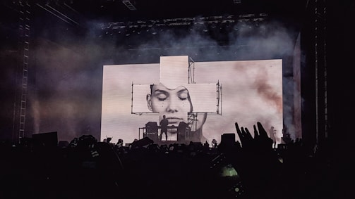 A concert stage is lit up with an impressive visual display featuring a large screen showing a black and white portrait of a person's face. Smoke or fog adds a dramatic effect to the atmosphere. Silhouettes of fans in the foreground raise their hands and capture the moment with their phones.