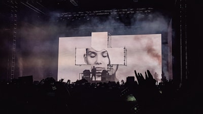A concert stage is lit up with an impressive visual display featuring a large screen showing a black and white portrait of a person's face. Smoke or fog adds a dramatic effect to the atmosphere. Silhouettes of fans in the foreground raise their hands and capture the moment with their phones.