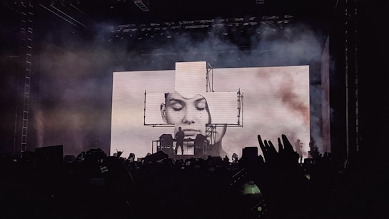 A concert stage is lit up with an impressive visual display featuring a large screen showing a black and white portrait of a person's face. Smoke or fog adds a dramatic effect to the atmosphere. Silhouettes of fans in the foreground raise their hands and capture the moment with their phones.