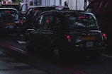 A dark street scene with several black taxis waiting in traffic. The image captures the vehicles from the rear with visible license plates. Reflections of trees can be seen on the car surfaces, and the brake lights are illuminated, casting a red glow. The atmosphere is dim, possibly indicating an overcast day or early evening.