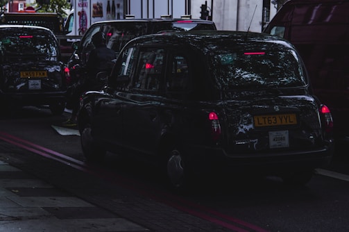 A dark street scene with several black taxis waiting in traffic. The image captures the vehicles from the rear with visible license plates. Reflections of trees can be seen on the car surfaces, and the brake lights are illuminated, casting a red glow. The atmosphere is dim, possibly indicating an overcast day or early evening.