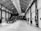 A spacious industrial-style interior with high ceilings and visible metal beams, filled with people engaged in viewing art or informational displays mounted on the walls. Some individuals are gathered around tables, possibly discussing or taking notes.