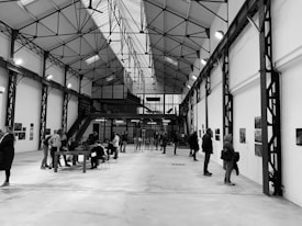 A spacious industrial-style interior with high ceilings and visible metal beams, filled with people engaged in viewing art or informational displays mounted on the walls. Some individuals are gathered around tables, possibly discussing or taking notes.