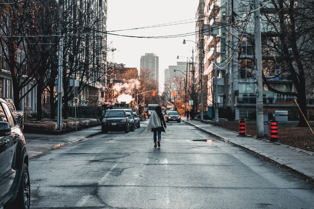 woman walking near vehicles parked on road between large buildings