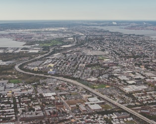 An aerial view of an urban area with a combination of residential, commercial, and industrial zones. The cityscape includes a network of roads and highways, and green areas scattered throughout the area. A body of water is visible in the distance, along with various buildings and infrastructure.