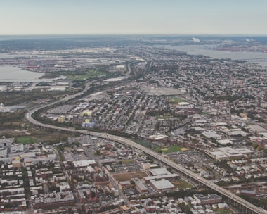 An aerial view of an urban area with a combination of residential, commercial, and industrial zones. The cityscape includes a network of roads and highways, and green areas scattered throughout the area. A body of water is visible in the distance, along with various buildings and infrastructure.