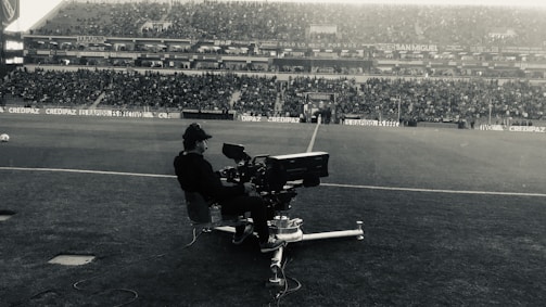 A person is operating a large camera on a dolly at the edge of a football field, with a crowded stadium in the background. The scene captures the atmosphere of a live sports event, and the audience is mostly seated, watching the action unfold.
